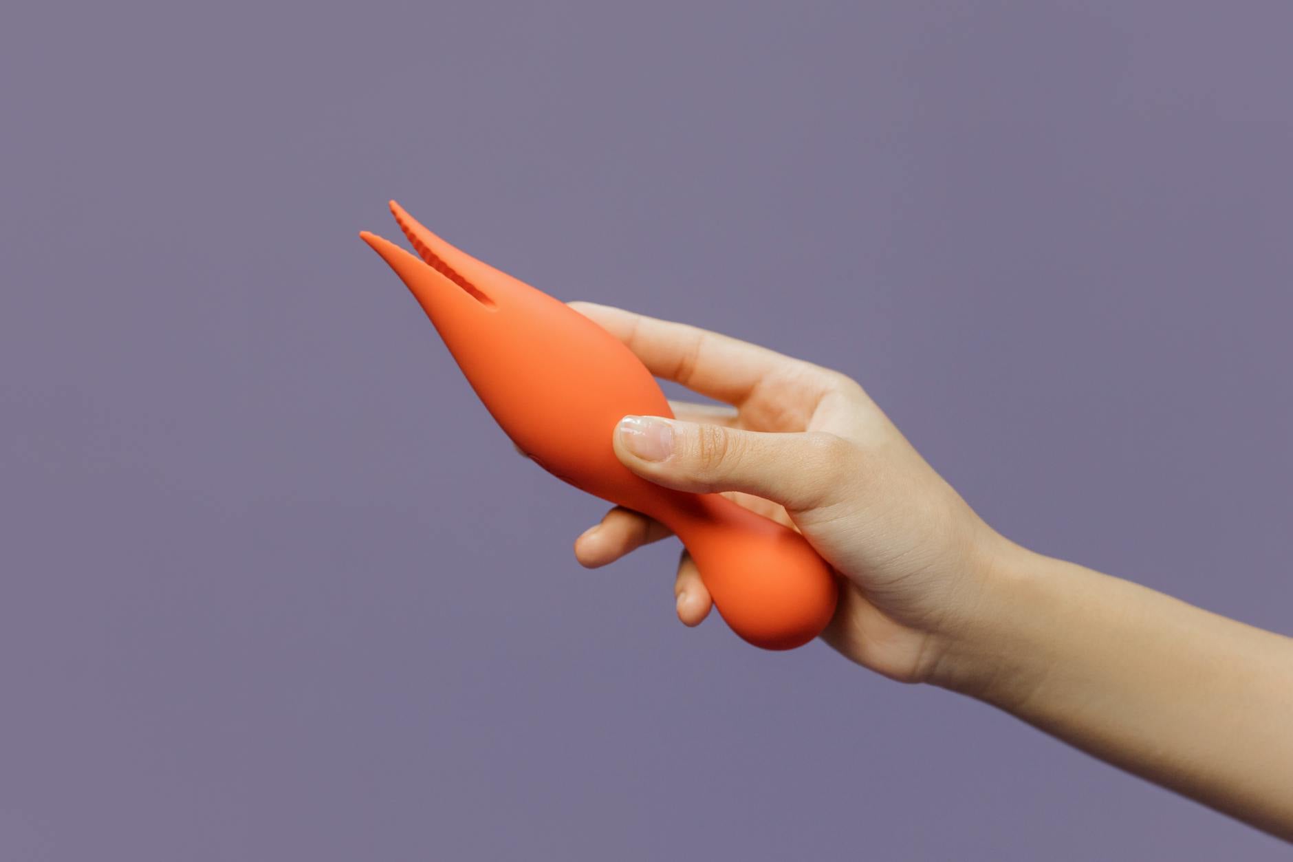 Mano sosteniendo vibrador Lemon naranja sobre fondo morado minimalista, mostrando sensualidad moderna